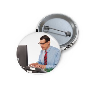 May include: Two round pinback buttons. One button features a man wearing glasses, a blue shirt, and a red striped tie, working on a computer. The other button is a silver metal back.