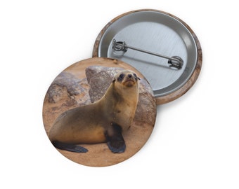 May include: A brown and gray seal pup with black eyes sits on a sandy beach with rocks in the background.