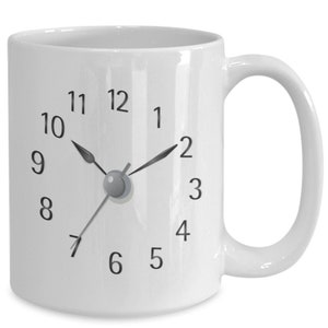 Könnte beinhalten: Weiße Keramik-Kaffeetasse mit einem Zifferblattmuster. Die Uhrzeiger sind auf 7:00 Uhr eingestellt.