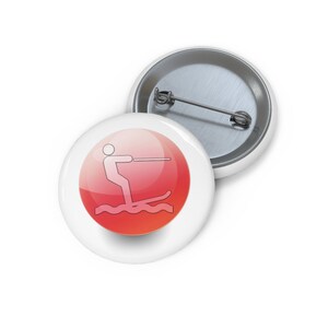 May include: A red and white button with a white silhouette of a person water skiing. The button is round and has a glossy finish.