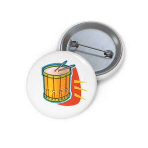 Op de afbeelding: Een witte knop met een cartoonillustratie van een gele trommel met rode en groene accenten. De trommel heeft twee drumsticks erboven en een rode en oranje achtergrond met gele stralen.