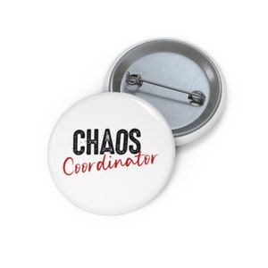 May include: White button with the words "CHAOS Coordinator" in black and red text. The button has a metal pin back. One button is in focus, and another is slightly out of focus in the background.
