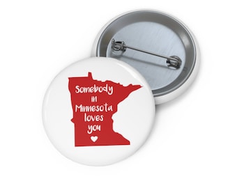 May include: White button with a red outline of the state of Minnesota. The text "Somebody in Minnesota loves you" is written in white on the red outline. A small white heart is below the text.