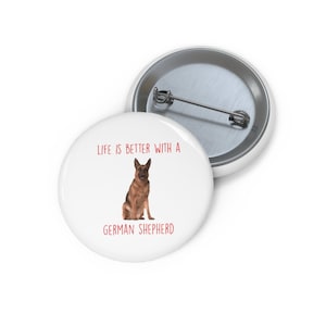 May include: White button with a red inscription that reads "Life is better with a German Shepherd". A brown and black German Shepherd dog is sitting in the center of the button.