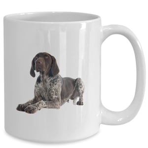 May include: White ceramic mug featuring a German Shorthaired Pointer dog. The dog is brown and white, lying down with its head up. The mug has a large handle and a glossy finish, perfect for coffee or tea.