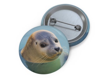 May include: A close-up of a seal's face peeking out of the water. The seal has a light gray coat and dark eyes. The background is a light blue.