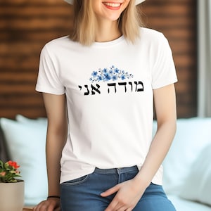 May include: White t-shirt with a blue floral design and the Hebrew text "מוֹדֶה אֲנִי" printed on the front.