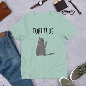 May include: A light green t-shirt with the word "Torttitude" printed in black. A cartoon cat with a grumpy expression is giving the middle finger.