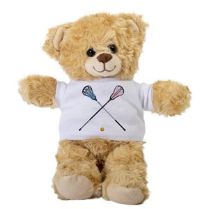 May include: A brown teddy bear wearing a white t-shirt with a graphic of two lacrosse sticks crossed over each other.