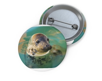 May include: A close-up of a seal's face peeking out of the water. The seal has brown fur and black eyes. The water is a turquoise color and the background is a blurry green.