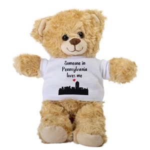 Könnte beinhalten: Ein hellbrauner Teddybär, der ein weißes T-Shirt mit dem Text "Jemand in Pennsylvania liebt mich" und eine schwarze Silhouette einer Stadtlandschaft mit einem roten Herzen darüber trägt.