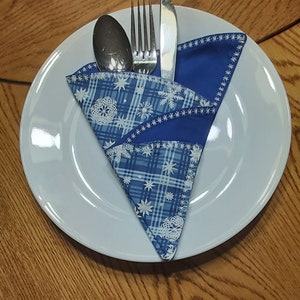 May include: A white plate with a blue and white patterned napkin folded into a triangular shape. A fork, knife, and spoon are placed inside the napkin.