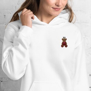 May include: White hoodie with a small embroidered teddy bear wearing a red bow tie. The text "XO" is embroidered above the bear. The hoodie has a drawstring hood and a front pocket.