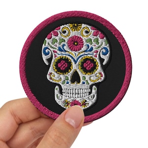 May include: Embroidered patch featuring a colorful sugar skull design. The skull is white with intricate floral and decorative patterns in pink, yellow, blue, and green. The patch has a black background with a magenta border.