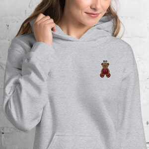 May include: A light gray hoodie with a front pocket and a drawstring hood. The hoodie features a small embroidered teddy bear with a red bow and the letters "XO" above it. The teddy bear is brown with dark brown accents.