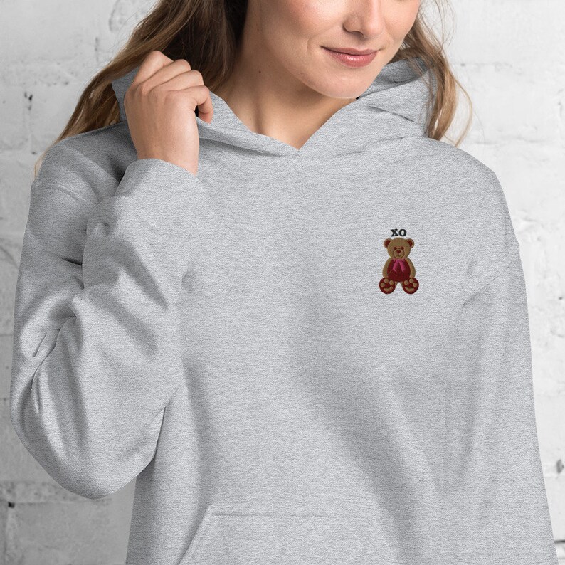 May include: A light gray hoodie with a front pocket and a drawstring hood. The hoodie features a small embroidered teddy bear with a red bow and the letters "XO" above it. The teddy bear is brown with dark brown accents.