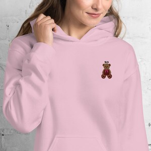 May include: A light pink hoodie with a front pocket and a drawstring hood. The hoodie features an embroidered teddy bear with a red bow and the letters "XO" above it. The person in the image is wearing the hoodie.
