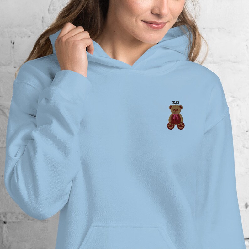 May include: Light blue hoodie with a small embroidered teddy bear wearing a red bow tie. The text "XO" is embroidered above the bear. The hoodie has a front pocket and a drawstring hood.