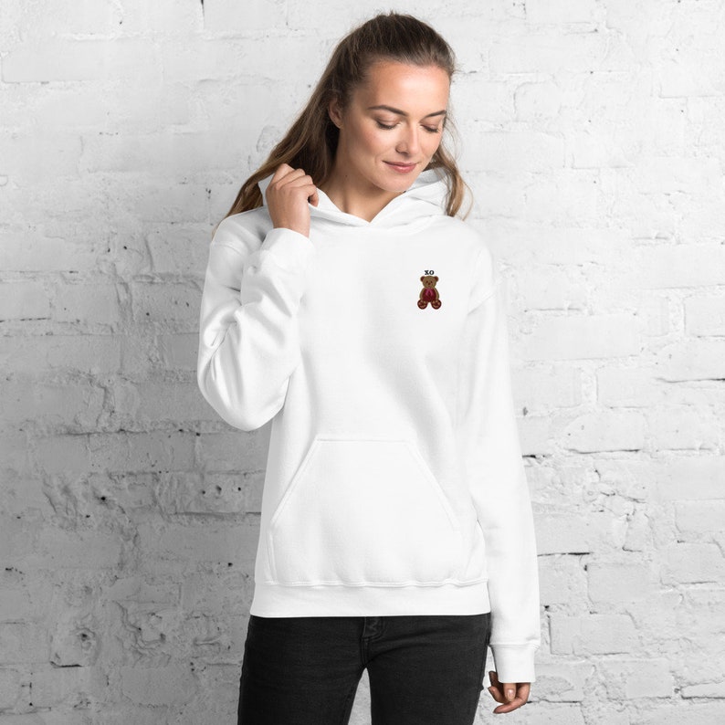 May include: White hoodie with a kangaroo pocket and a drawstring hood. The hoodie features a small embroidered teddy bear design with the letters "XO" above it. The teddy bear is brown with a red heart.