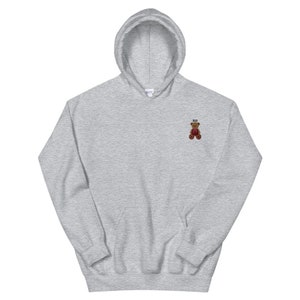May include: A light gray hoodie with a drawstring hood and a kangaroo pocket. The hoodie features a small embroidered teddy bear with the letters "XO" above it. The teddy bear is brown with red accents.