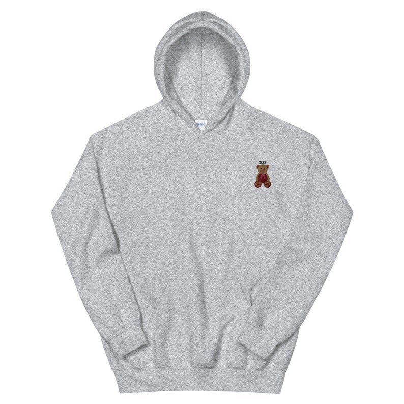 May include: A light gray hoodie with a drawstring hood and a kangaroo pocket. The hoodie features a small embroidered teddy bear with the letters "XO" above it. The teddy bear is brown with red accents.