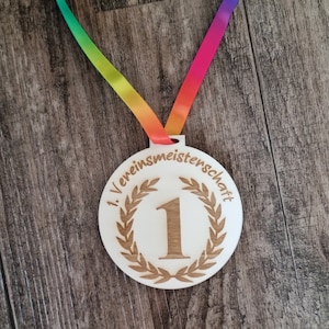 Könnte beinhalten: Eine Holzmedaille mit einem Regenbogenband. Die Medaille hat ein Lorbeerkranz-Design und die Zahl "1" in der Mitte. Der Text "1. Vereinsmeisterschaft" ist um den Rand der Medaille graviert.