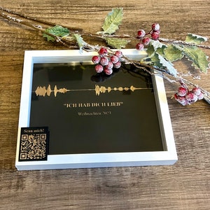 Könnte beinhalten: Ein gerahmter schwarzer Druck mit goldenem Text und einem Wellenform-Design. Der Text lautet "ICH HAB DICH LIEB" und "Weihnachten 2023". Ein QR-Code-Aufkleber befindet sich in der unteren linken Ecke. Der Rahmen ist weiß, dekoriert mit Beeren.