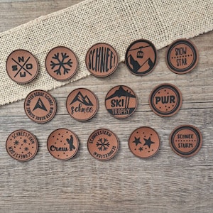Könnte beinhalten: Eine Sammlung brauner Lederpatches mit schwarzen Grafiken und Texten zum Thema Wintersport. Die runden Patches zeigen Schneeflocken, Berge, Skilifte und das Wort "Schnee". Die Patches sind in verschiedenen Größen erhältlich.
