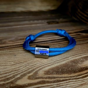 Könnte beinhalten: Ein blaues Paracord-Armband mit einer silbernen Metallperle, die mit "NOTFALLNUMMER 0160 Mama" graviert ist.