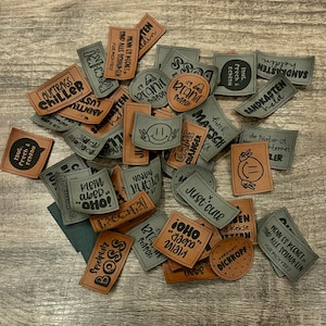 Könnte beinhalten: Eine Sammlung von braunen und grauen Stoff-Patches mit verschiedenen Sprüchen und Designs, die darauf gedruckt sind. Die Patches beinhalten Sätze wie "Game Changer", "Just Cute", "Sandkasten", "It's Branie Season", "Auftrags Chiller", "Klein aber Oho!" und "Spielplatz Boss".