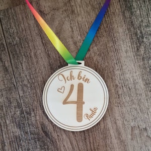 Könnte beinhalten: Eine Holzmedaille mit einem Regenbogenband. Die Medaille trägt die Gravur "Ich bin 4 Paula".