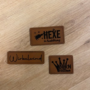 Könnte beinhalten: Drei braune Lederpatches mit schwarzem Text. Der erste Patch sagt "HEXE in Ausbildung" mit einem Besen und Sternen. Der zweite Patch sagt "Wirbelwind". Der dritte Patch sagt "ZORN Froschen" mit einer Krone.