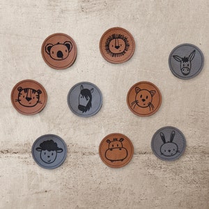 Könnte beinhalten: Neun runde braune und graue Filzpatches mit schwarzen Strichzeichnungen verschiedener Tiere. Die Tiere umfassen einen Koala, einen Löwen, einen Esel, einen Tiger, ein Pferd, eine Katze, ein Flusspferd, ein Kaninchen und ein Schaf.