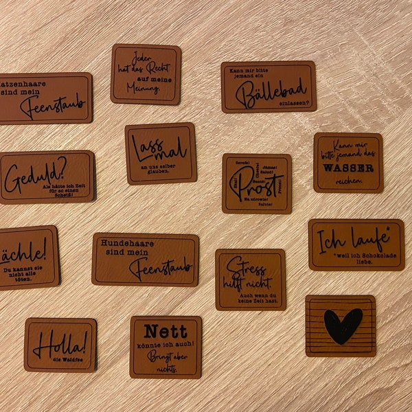 May include: A collection of 12 brown leather coasters with various sayings printed in black. The sayings include "Katzenhaare sind mein Feenstaub", "Geduld?", "Lächle!", "Jeder hat das Recht auf meine Meinung", "Lass mal", "Hundehaare sind mein Feenstaub", "Kann mir bitte jemand ein Bällebad einräumen?", "Stress hilft nicht", "Nett könnte ich auch! Bringt aber nichts", "Kann mir bitte jemand das Wasser reichen", "Ich laufe *weil ich Schokolade liebe*" and "Holla! die Waldfee".