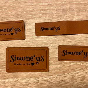 Könnte beinhalten: Vier braune Lederetiketten mit dem Text "Simone's" in schwarzer Schrift. Zwei der Etiketten enthalten auch den Text "Made with" und ein kleines Herzsymbol.