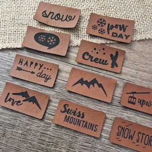 Könnte beinhalten: Zehn braune Lederpatches mit schwarzem Text und Designs. Die Patches zeigen Sätze wie "Schnee", "Powder Day", "Happy... Day", "Crew", "Liebe", "Schweizer Berge", "Schneesturm" und "Uper".