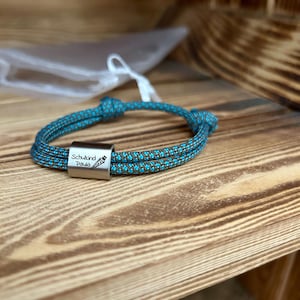 Könnte beinhalten: Ein blaues und braunes geflochtenes Seil-Armband mit einem silbernen Anhänger, auf dem "Schulkind Paula" mit einer kleinen Zeichnung einer Karotte steht.