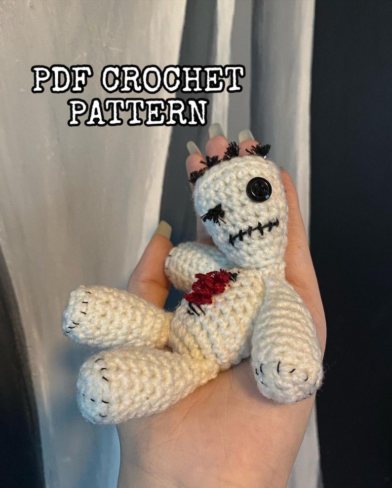 Puede incluir: Una mu&ntilde;eca vud&uacute; de crochet blanco con ojos negros, boca cosida y un coraz&oacute;n rojo. La mu&ntilde;eca tiene un cuerpo cosido y le faltan algunas puntadas. La mu&ntilde;eca est&aacute; sostenida en una mano con u&ntilde;as largas pintadas.
