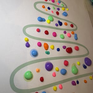 Puede incluir: Un papel blanco con un contorno verde de un árbol de Navidad. El árbol está decorado con puntos de pintura de colores en varios tonos de rojo, naranja, amarillo, verde, azul, morado y rosa.