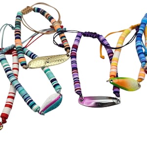 May include: A collection of five adjustable bracelets featuring colorful heishi clay beads in various striped patterns. Each bracelet includes a central metallic or glitter-finished oval charm, with adjustable cord closures for a secure fit.