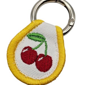 May include: A cherry-themed keychain with a silver clasp. The embroidered patch features two red cherries with green leaves on a white background, framed by a yellow border. A fun accessory.