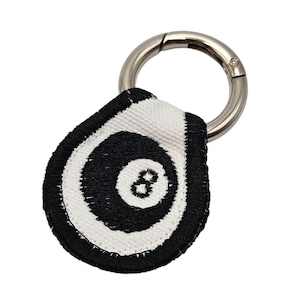 May include: A black and white 8-ball keychain with a silver metal clasp. The keychain features a teardrop-shaped design with a black border and a white and black 8-ball graphic. The clasp is a silver metal ring.