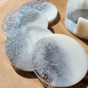 May include: Set of four white and silver glitter coasters with a matching larger coaster. The coasters are round and have a white base with silver glitter embedded in the resin.
