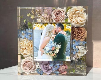 May include: A square clear resin photo frame featuring embedded dried flowers in shades of dusty rose, cream, and light blue. The frame surrounds a central photograph of a couple in wedding attire, with the flowers arranged along the border.