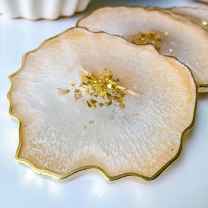 May include: Set of four resin coasters with a gold trim. The coasters have a creamy white and gold color scheme with gold flakes embedded in the resin. The edges have a wavy, organic shape.