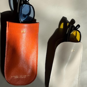 Könnte beinhalten: Zwei vertikale Lederetuis für Brillen, eines in gebranntem Orange und eines in Off-White. Jedes Etui ist unten mit goldgeprägten Initialen und einem Markennamen versehen. Das orangefarbene Etui enthält eine Brille mit schwarzem Rahmen, das weiße Etui eine Brille mit gelb getönten Gläsern.