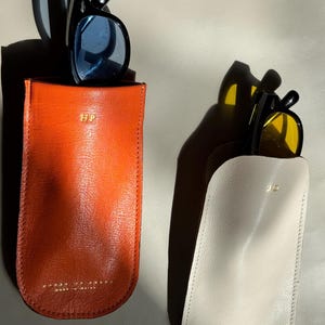 May include: Two leather eyeglass cases, one orange and one off-white, each holding sunglasses. The orange case has blue-tinted lenses, and the off-white case has yellow-tinted lenses. Both cases have gold lettering and a stitched edge.