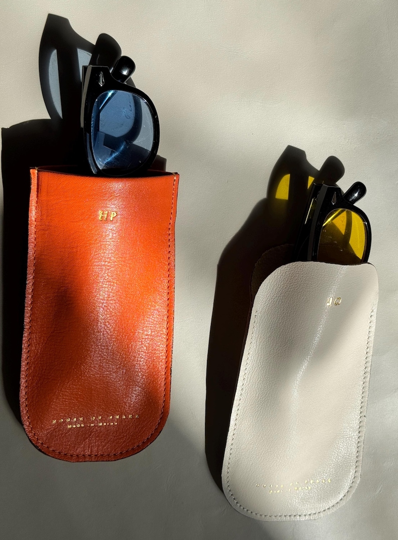 May include: Two leather eyeglass cases, one orange and one off-white, each holding sunglasses. The orange case has blue-tinted lenses, and the off-white case has yellow-tinted lenses. Both cases have gold lettering and a stitched edge.