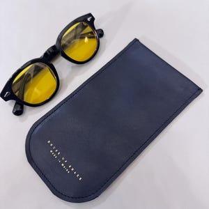May include: Black-framed sunglasses with yellow lenses rest beside a navy blue leather glasses case. The case has rounded edges and gold lettering that reads "HOUSE OF SNAKE Made in Maine."