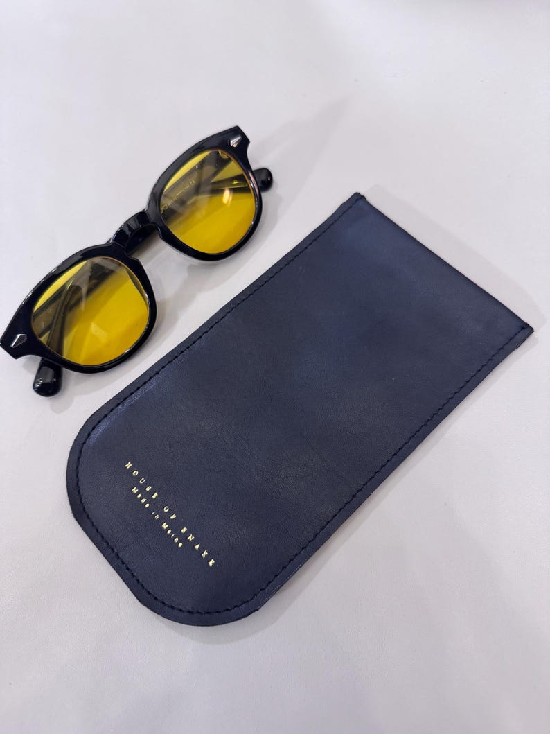 May include: Black-framed sunglasses with yellow lenses rest beside a navy blue leather glasses case. The case has rounded edges and gold lettering that reads "HOUSE OF SNAKE Made in Maine."